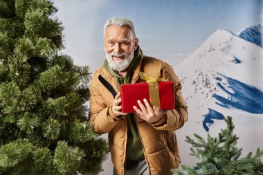Beyaz sakallı neşeli Noel Baba çam ağacının yanında duruyor ve kışın kameraya gülümsüyor.