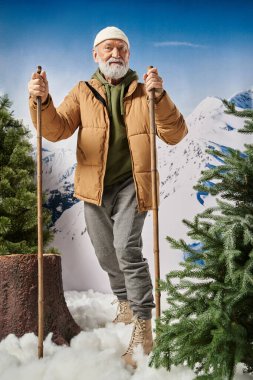 Ağaçların yanında elinde kayak sopalarıyla yakışıklı sporcu Noel Baba, kış konsepti.
