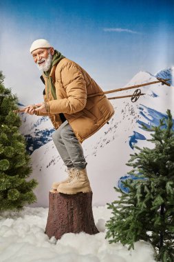 Beyaz sakallı Noel Baba, ağaç kütüğüne çömelmiş kayak sopalarıyla kameraya gülümsüyor, kış konsepti.