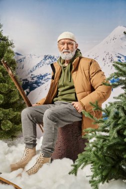 Noel Baba gibi giyinmiş bir adam ağaç kütüğünde oturuyor ve elinde dağ zeminli kayak sopaları tutuyor.