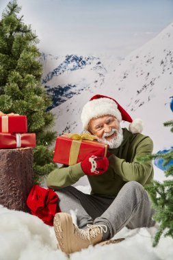 Noel Baba gibi giyinmiş neşeli bir adam hediye tutuyor ve ağaç kütüğünün yanındaki karda oturuyor, kış konsepti.