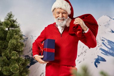 cheerful man in Santa costume holding present bag and big gift looking at camera, winter concept clipart