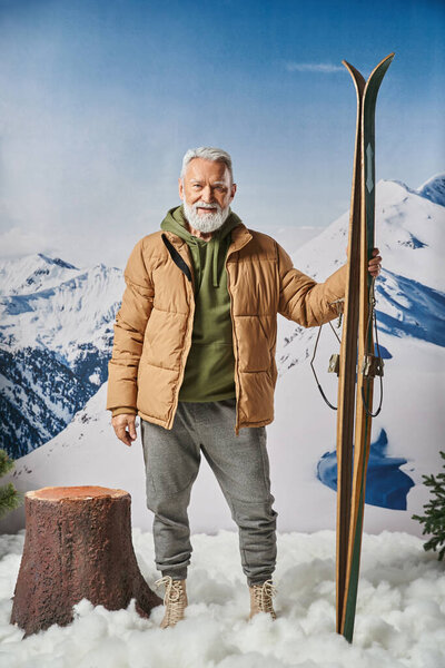 sporty cheerful Santa with white beard holding ski with snowy mountains on backdrop, winter concept