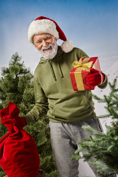 Радостный Санта держит в руках подарочную сумку и красный подарок, улыбаясь в камеру, зимняя концепция
