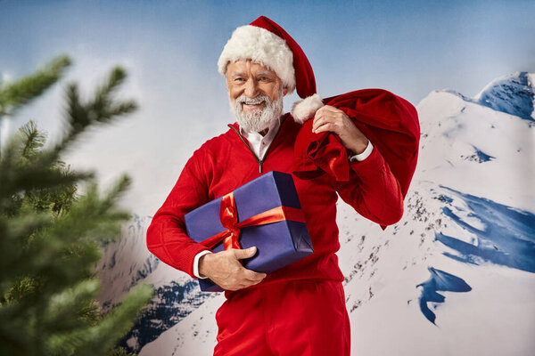 радостный Санта в красной одежде улыбаясь в камеру с подарком и подарочной сумкой в руках, зимняя концепция