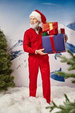 Gülümseyen sakallı Noel Baba elinde hediyelerle dağların arkasına bakıyor, kış konsepti.