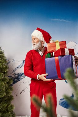 Yakışıklı Noel Baba çam ağaçlarının yanında hediyeler tutuyor. Başka tarafa bakıyor ve gülümsüyor. Kış konsepti.