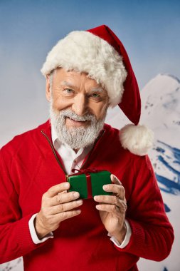 Beyaz sakallı neşeli Noel Baba küçük bir hediye tutuyor ve kameraya gülümsüyor, kış konsepti.