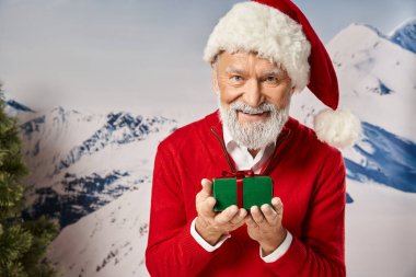 Neşeli, beyaz sakallı Noel Baba avuçlarında gülümsüyor ve onu kameraya gösteriyor, kış konsepti.