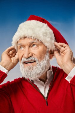 Noel Baba kostümü giymiş neşeli bir adamın portresi Noel şapkasına dokunuyor, uzaklara bakıyor, kış konsepti.