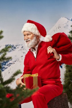Neşeli Noel Baba ağaç kütüğünde hediye çantasıyla oturuyor ve uzaklara bakıyor, kış konsepti.