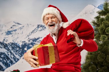 Noel Baba gibi giyinmiş neşeli bir adam kameraya mutlu bir şekilde gülümsüyor. Hediye çantası ve hediye, kış konsepti.