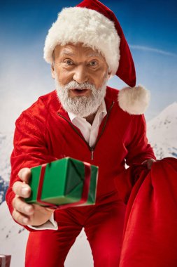Beyaz sakallı Noel Baba kırmızı kurdeleyle hediye veriyor ve kameraya bakıyor, kış konsepti.