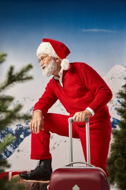 Gözlüklü beyaz sakallı neşeli Noel Baba ağaç kütüğünün yanında bavulla poz veriyor, kış konsepti.