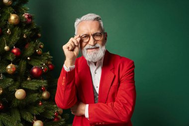 Noel Baba gibi giyinmiş şık bir adam kollarını kavuşturup köknar ağacının yanında gözlük takıyor.