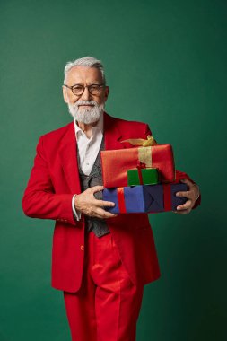 Zarif Noel Baba gibi giyinmiş yakışıklı bir adam koyu yeşil arka planda hediyelerle poz veriyor, kış konsepti.