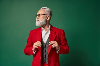 good looking elegant Santa with glasses touching his suit and looking away, winter concept clipart