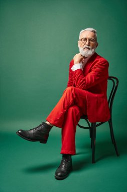 Noel Baba gibi giyinmiş, gözlüklü şık bir adam sandalyeye oturmuş, eli yüzüne yakın, kış konsepti.