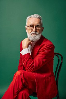 Kırmızı takım elbiseli şık, yakışıklı Noel Baba eli yüzü yakınken sandalyede poz veriyor. Noel konsepti.