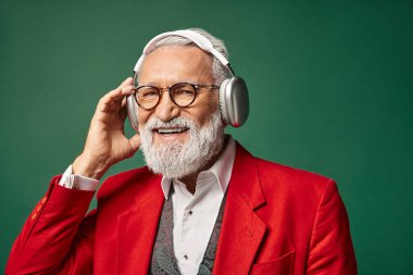 Kırmızı Noel Baba kostümlü şık bir adam kulaklık takıyor ve kameraya gülümsüyor, kış konsepti.