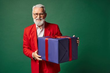 Sakallı ve gözlüklü zarif Noel Baba kocaman bir hediye tutuyor ve içten gülümsüyor, kış konsepti.
