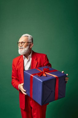 Şık Noel Baba yeşil arka planda büyük bir hediye tutuyor ve uzaklara bakıyor, kış konsepti.