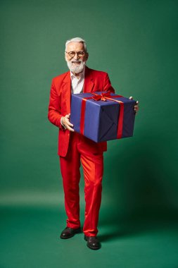 Noel Baba gibi giyinmiş yakışıklı bir adam yeşil zeminde büyük bir hediyeyle poz veriyor, kış konsepti.