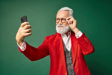 Zarif Noel Baba gibi giyinmiş şık yakışıklı adam telefonda ciddi selfie çekiyor, kış konsepti.