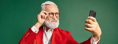 Kırmızı Noel Baba kostümlü şık sakallı adam telefonda ciddi selfie çekiyor, kış konsepti, afiş.