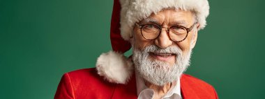 Kırmızı şapkalı ve gözlüklü zarif Noel Baba kameraya, kış konseptine, afişe neşeyle gülümsüyor.