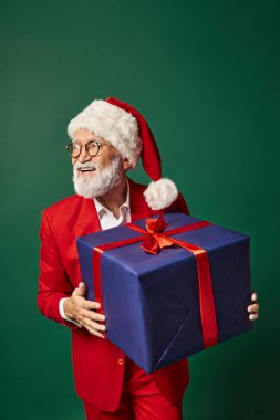 Noel Baba gibi giyinmiş neşeli bir adam elinde kocaman bir hediye, gülümsüyor ve uzaklara bakıyor, Noel konsepti.