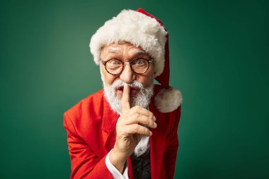 stylish Santa in red hat and glasses showing silence gesture looking at camera, winter concept clipart