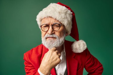 Noel Baba gibi giyinmiş, eli çenesinin altında kameraya bakan ciddi bir adam, kış konsepti.