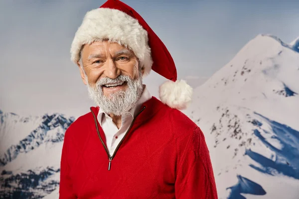 stock image good looking jolly Santa in red attire smiling at camera with mountain backdrop, winter concept
