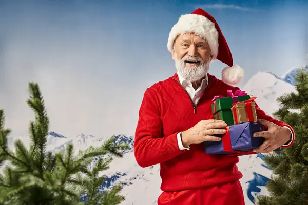 Beyaz sakallı neşeli Noel Baba karlı dağ zeminli hediye yığınını tutuyor, kış konsepti