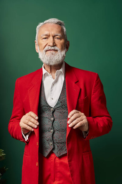 elegant white bearded Santa posing with hands on suit and looking at camera, winter concept
