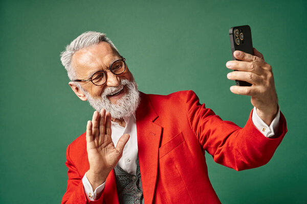 stylish jolly Santa with beard and glasses gesturing and waving at phone camera, winter concept