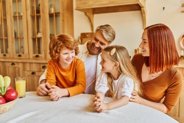 joyful parents with daughter and son sitting near fresh fruits and orange juice in cozy kitchen clipart