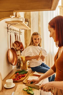 happy girl sitting with sandwich on kitchen counter near smiling mother preparing breakfast clipart