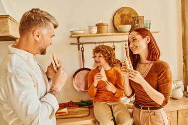 Neşeli ebeveynler ve mutlu oğul kahvaltıda sandviç yiyorlar. Modern mutfakta, aile yemeklerinde.