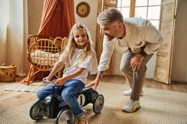 cheerful man assisting cute daughter riding toy car in modern living room at home, playing together clipart