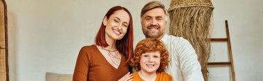 smiling parents with redhead son sitting looking at camera in modern bedroom, horizontal banner clipart