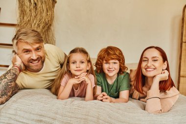 smiling parents with son and daughter lying ibn cozy bedroom and looking at camera, bonding moments clipart