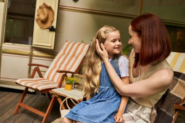 smiling mother stroking hair of cute daughter while sitting next to trailer home, love and care clipart