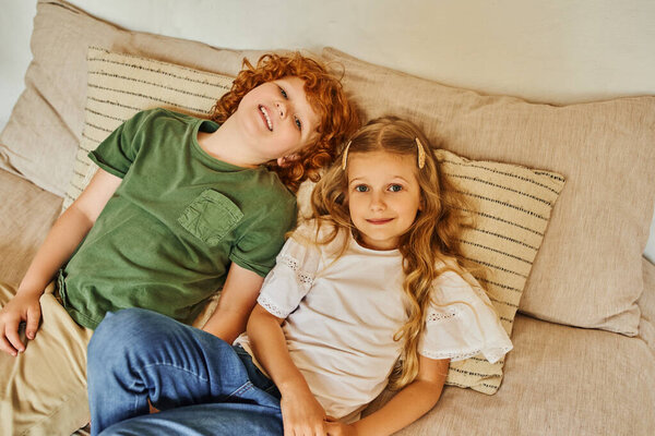 top view of brother and sister lying on bed with soft pillows and looking at camera, happy moments