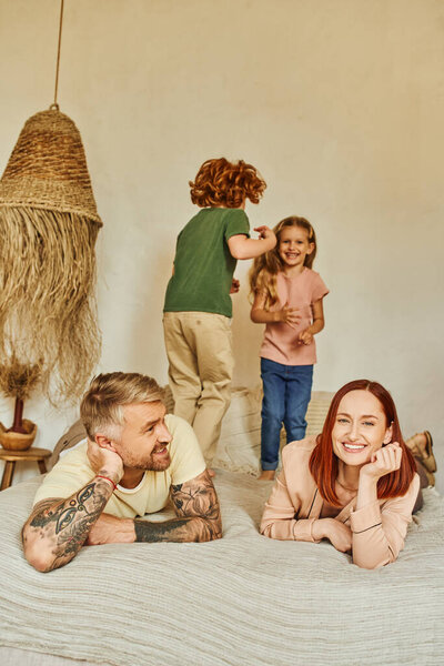 счастливый муж и жена улыбаются рядом взволнованных детей, веселящихся на кровати дома, время релаксации