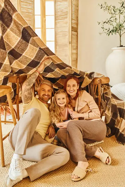 stock image excited parents with happy daughter sitting under blanket hut in living room, playing together