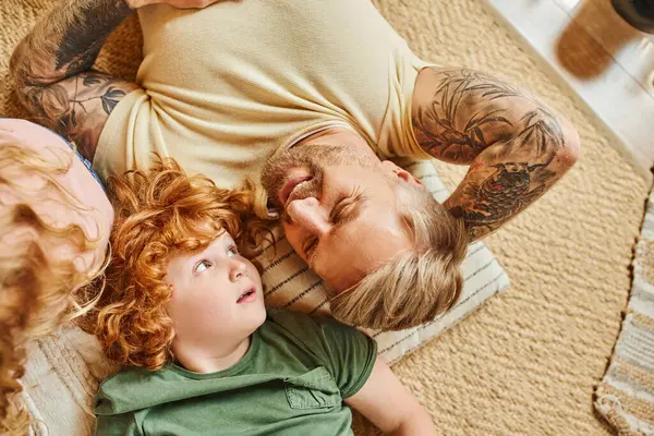 stock image top view of cheerful tattooed man lying down with redhead son on floor in living room, bonding