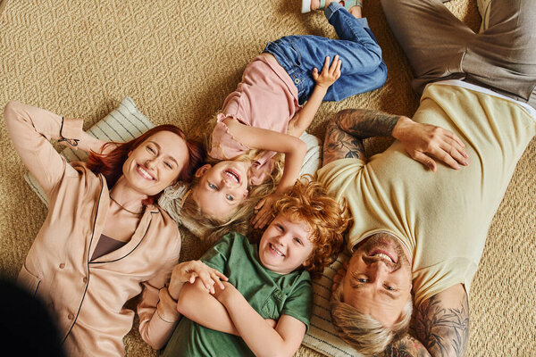 top view of smiling parents and children lying down on floor in living room and smiling at camera