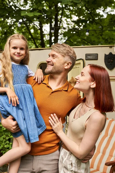 joyful man holding cute daughter near happy wife and mobile home in trailer park, leisure together